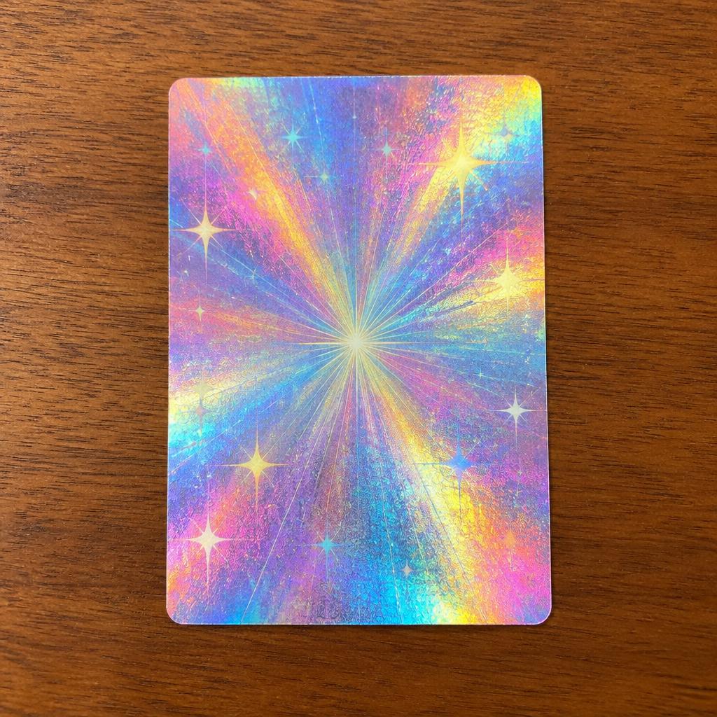 The back of one of the trading cards — a shiny holographic rainbow foil back with a sparkly starburst pattern, lying on a wooden table